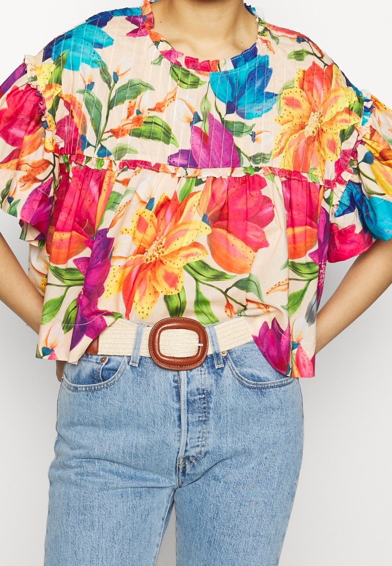 Blomstret bluse med flæseærmer, prydet med flerfarvede liljer og blade på en lys baggrund, sat sammen med lyseblå jeans og et beige bælte.