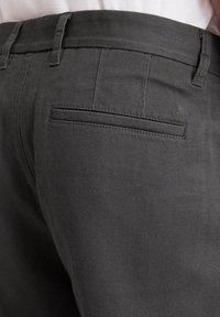 Graue Hose mit glatter Textur, ausgestattet mit einer Gesäßtasche und Gürtelschlaufen. Dezenter Markenakzent in der Nähe der Tasche als Detail.