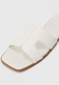 Sandale albe din material neted, cu un design deschis, prezentând două barete curbate și o talpă plată din cauciuc maro.