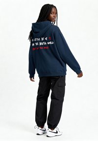 Felpa blu con cappuccio, con testo bianco e rosso sul retro, abbinata a pantaloni cargo neri e sneakers bianche. Vestibilità comoda, texture morbida.