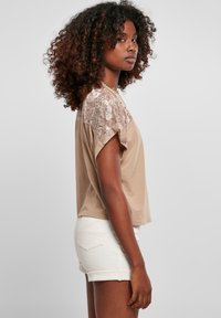 Beige kortærmet top med blonder detaljer på skuldrene og en afslappet pasform. Kombinationen inkluderer hvide denimshorts. Den teksturerede blonde detaljer er fremtrædende.