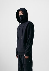 Schwarzer Kapuzenpullover mit abnehmbarem Gesichtsschutz, mit Kordelzügen und entspannter Passform. Der Stoff wirkt weich mit einer matten Textur.