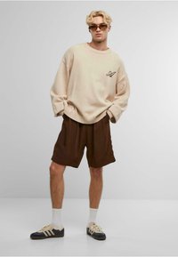 Pulover oversized de culoare bej deschis cu un logo marron, combinat cu pantaloni scurți de culoare maro închis. Modelul poartă adidași bej și bleumarin, cu șosete albe.