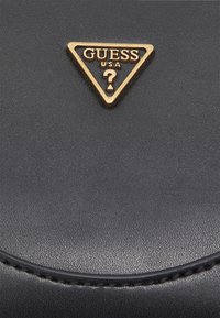 Černá kožená kabelka s výrazným zlatým logem ve tvaru trojúhelníku s textem "GUESS USA" a otazníkem. Viditelný texturovaný povrch.