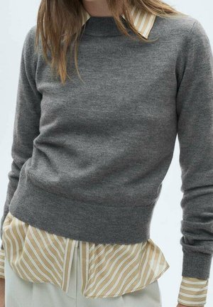Persona con un suéter gris sobre una camisa de botones a rayas amarillas y blancas, con pantalones blancos, mostrada desde los hombros hasta las caderas.