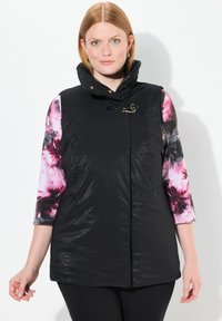 Schwarze ärmellose Weste mit hohem Kragen, glänzender Textur und goldenen Beschlägen. Die Innenärmel haben ein Pink- und Schwarz-Tie-Dye-Muster.