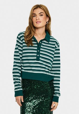 Woman with loose light brown hair wearing green and white striped long-sleeve collared shirt and green sequined skirt, standing against white background.