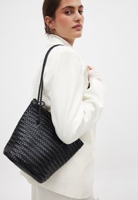 Mustan kudotun ämpärilaukun, jossa on teksturoitu pinta ja punottu kahva, pitää henkilö, joka on pukeutunut valkoiseen bleiseriin.