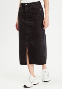 Jupe midi en denim noir avec une fente sur le devant, un design à cinq poches et des coutures discrètes. Associée à des baskets blanches.