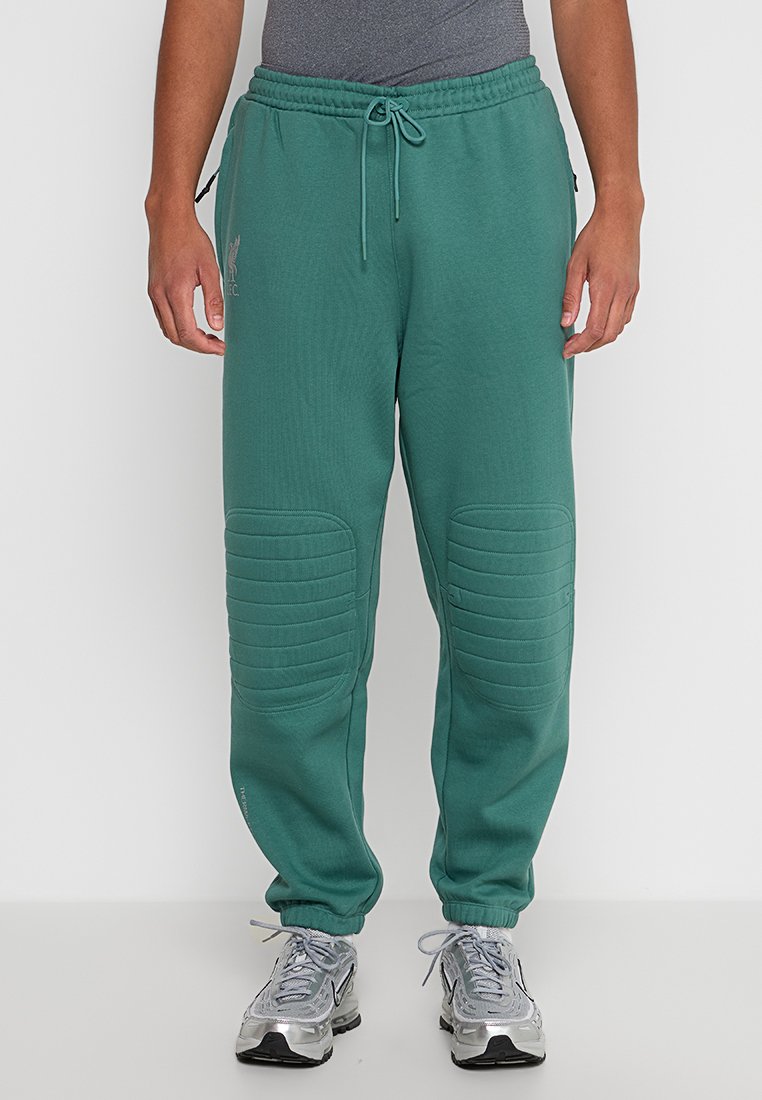 Uomo che indossa pantaloni jogging imbottiti turchesi e scarpe da corsa argentate, in piedi contro uno sfondo bianco uniforme.