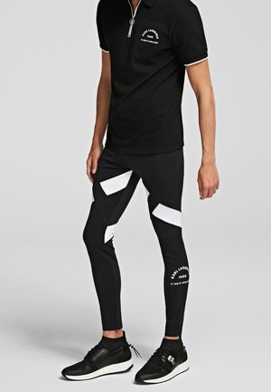 Hombre con polo negro con cremallera y leggings negros con patrones geométricos blancos y texto, combinado con zapatillas negras con suelas blancas.