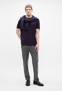 Σκούρο μωβ μπλούζα με λογότυπο, γκρι κομμένα παντελόνια, και μαύρα παπούτσια; ναυτικό πουλόβερ ριγμένο στους ώμους; μινιμαλιστικό φόντο.