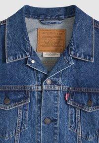 Džínová bunda v tmavomodrej farbe s golierom, náprsnými vreckami, mosadznými gombíkmi a koženou štítkou vo vnútri. Klasický dizajn.