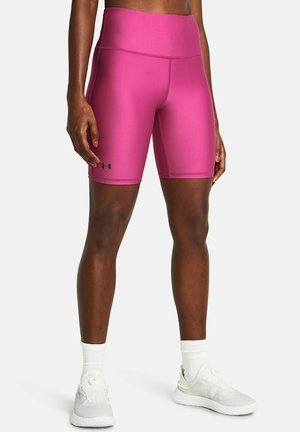 Atleet draagt felroze Under Armour compressieshorts, witte sokken en witte hardloopschoenen, staand tegen een effen achtergrond.