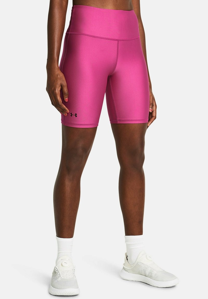 Athlète portant un short de compression Under Armour rose vif, des chaussettes blanches et des chaussures de course blanches, debout devant un fond uni.