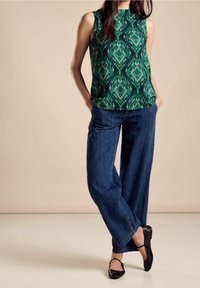 Ärmlös grön topp med ett geometriskt mönster, matchad med löst sittande blå jeans och svarta platta skor. Enkel och avslappnad outfit.