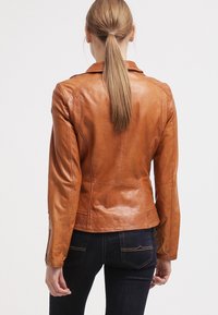 Giacca di pelle marrone con un design aderente, caratterizzata da una texture liscia, cuciture sagomate e un colletto modesto. Indossata sopra dei jeans scuri.