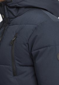 Granatowa pikowana kurtka z teksturowanym wykończeniem, kieszenią na piersi na suwak, regulowanym kapturem ze sznurkiem oraz okrągłym logo na rękawie.