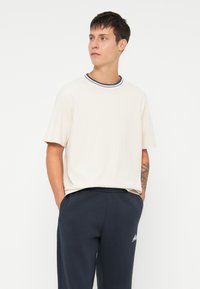Kortermet beige T-skjorte med en rund navy og hvit stripete krage, kombinert med mørk marineblå bukser, stående mot en ensfarget hvit bakgrunn.