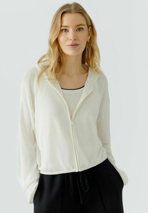 Femme blonde aux cheveux lâchés porte un sweat à capuche blanc zippé sur un haut clair et un pantalon noir, debout les mains dans les poches.