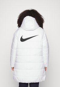 Biała puchowa kurtka z kapturem, z dużym czarnym logo Nike na plecach, pikowanym wzorem oraz elastycznym dołem i mankietami.