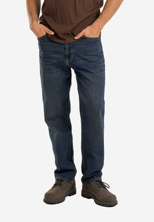 Mann trägt dunkelblaue Jeans, braune Schnürstiefel, mit Händen in den Taschen und teilweise sichtbarem braunen Hemd.