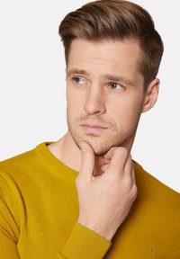 Musztardowa żółta sweter z dzianiny o okrągłym dekolcie i ściągaczami. Model dotykający podbródka, prezentujący teksturowany materiał i subtelny detal z logo.