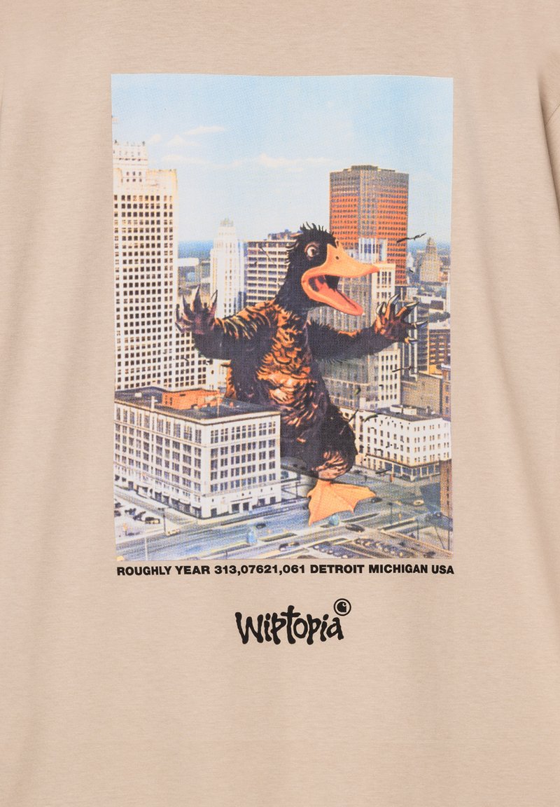 Camiseta beige con un gráfico colorido de una criatura gigante en un paisaje urbano. El texto en la parte inferior dice "Wliitopia" y las coordenadas de la ciudad.