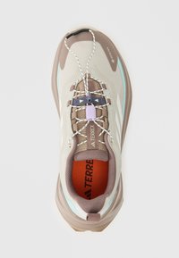 Beige en bruine sportschoen met een gestructureerde bovenkant, reflecterende accenten en elastische veters. De binnenkant heeft een feloranje inlegzool.
