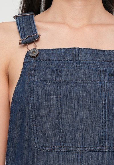 Salopete din denim de culoare bleumarin închis, cu bretele metalice ajustabile. Prezintă un buzunar frontal și un cusut vizibil. Materialul are o textură netedă.