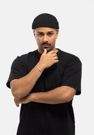 Berretto a coste nero, t-shirt nera, modello maschile con barba corta, mani incrociate sotto il mento, braccialetto argento al polso sinistro. Sfondo bianco.