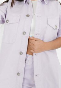 Lila skjorta med knappar i denim, med korta ärmar, två bröstfickor, silverknappar och en avslappnad passform.