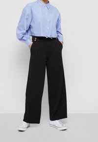 Blue and white striped button-up shirt with a tied hem, paired with black wide-leg trousers featuring gold buttons and white sneakers.