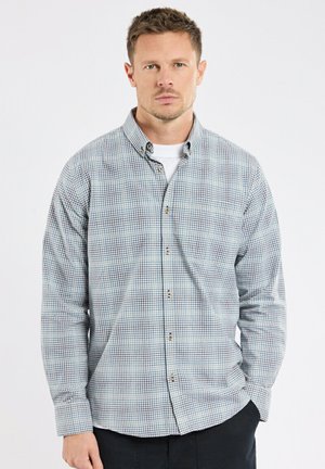 Homme portant une chemise à manches longues à carreaux bleus et blancs boutonnée par-dessus un t-shirt blanc, debout avec une main dans la poche sur un fond blanc.