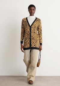 Kardigan s leoprintom v rumeni in črni barvi, z globokim V-izrezom, gumbom za zapiranje in sprednjimi žepi, usklajen z bež hlačami in dodatki.