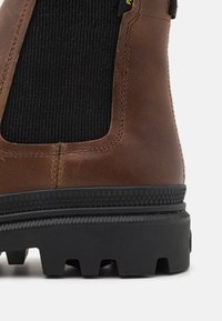 Botte Chelsea en cuir marron avec élastique noir côtelé, semelle en caoutchouc noir texturé et détails de couture visibles. Conçue pour la durabilité et le confort.