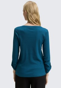 Vrouw met blond haar draagt een te groen langetermijnshirt met knoop-tab mouwen en zwarte broek, van achteren afgebeeld op een effen achtergrond.