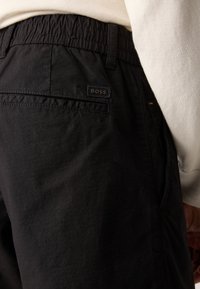 Gros plan sur un pantalon noir à taille élastique avec le logo BOSS près de la poche arrière, porté avec un haut beige à manches longues.