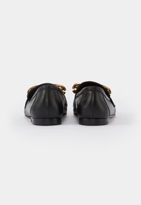 Mocassins en cuir noir à talons bas, vus de dos, avec un détail de chaîne dorée sur le dessus, sur fond blanc uni.