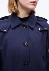 Γυναίκα με σκούρο μπλε παλτό με κουμπιά και φαρδιά γιακά, φορά χρυσό στεφάνι ως σκουλαρίκι, σε απλό λευκό φόντο.