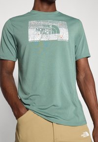 Zielona koszulka z krótkim rękawem, wykonana z miękkiego materiału, z białym nadrukiem topograficznym i logo "THE NORTH FACE" oraz napisem "NEVER STOP EXPLORING."