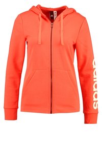 Sweat à capuche zippé corail en tissu doux, avec une capuche à cordon, deux poches avant et un grand logo blanc le long de la manche gauche.