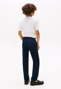 Copil cu păr creț, văzut de spate, purtând un tricou alb simplu cu mâneci scurte, pantaloni bleumarin și pantofi negri pe un fundal alb.