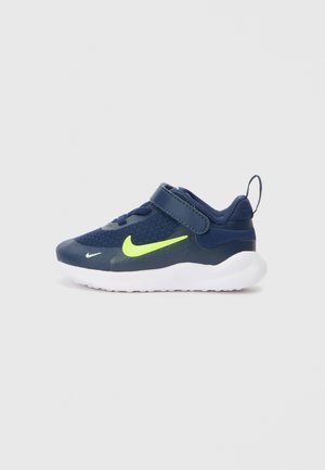 Baskets bleu marine avec dessus en mesh, swoosh jaune néon, sangle réglable et semelle blanche. Comprend une languette arrière pour un enfilage facile.