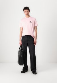 T-shirt en coton rose avec un petit logo, jeans noirs à jambe large et sac fourre-tout en cuir noir. Le modèle porte des chaussures noires, debout sur un fond blanc.