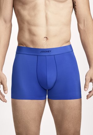Männlicher Oberkörper, der blaue Boxershorts der Marke Jockey trägt, mit einem definierten Bauch und teilweise sichtbaren Armen mit Tätowierung am rechten Arm.