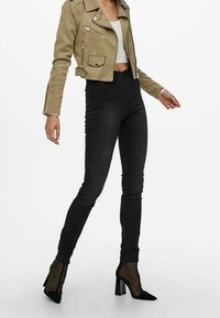 Person som bär en tanfärgad, kort jacka, vit crop top, högmidjade svarta skinny jeans och svarta nätade högklackade ankelskor.