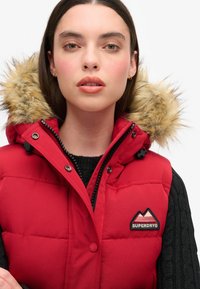 Rote Steppweste mit einer Fellkapuze, ausgestattet mit einem Reißverschluss und Druckknopfverschluss, schwarzen Akzenten und einem Berglogo auf der Brust.