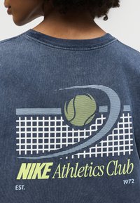Άτομο που φοράει μπλε μπλουζάκι Nike με γραφικό από μπάλα του τένις, σχέδιο με δίχτυ και κείμενο "Nike Athletics Club Est. 1972" στην πλάτη.