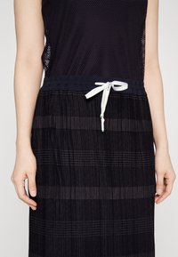 Femme portant un haut en résille noire et une jupe plissée noire à rayures texturées horizontales, attachée avec une ceinture à cordon blanche.